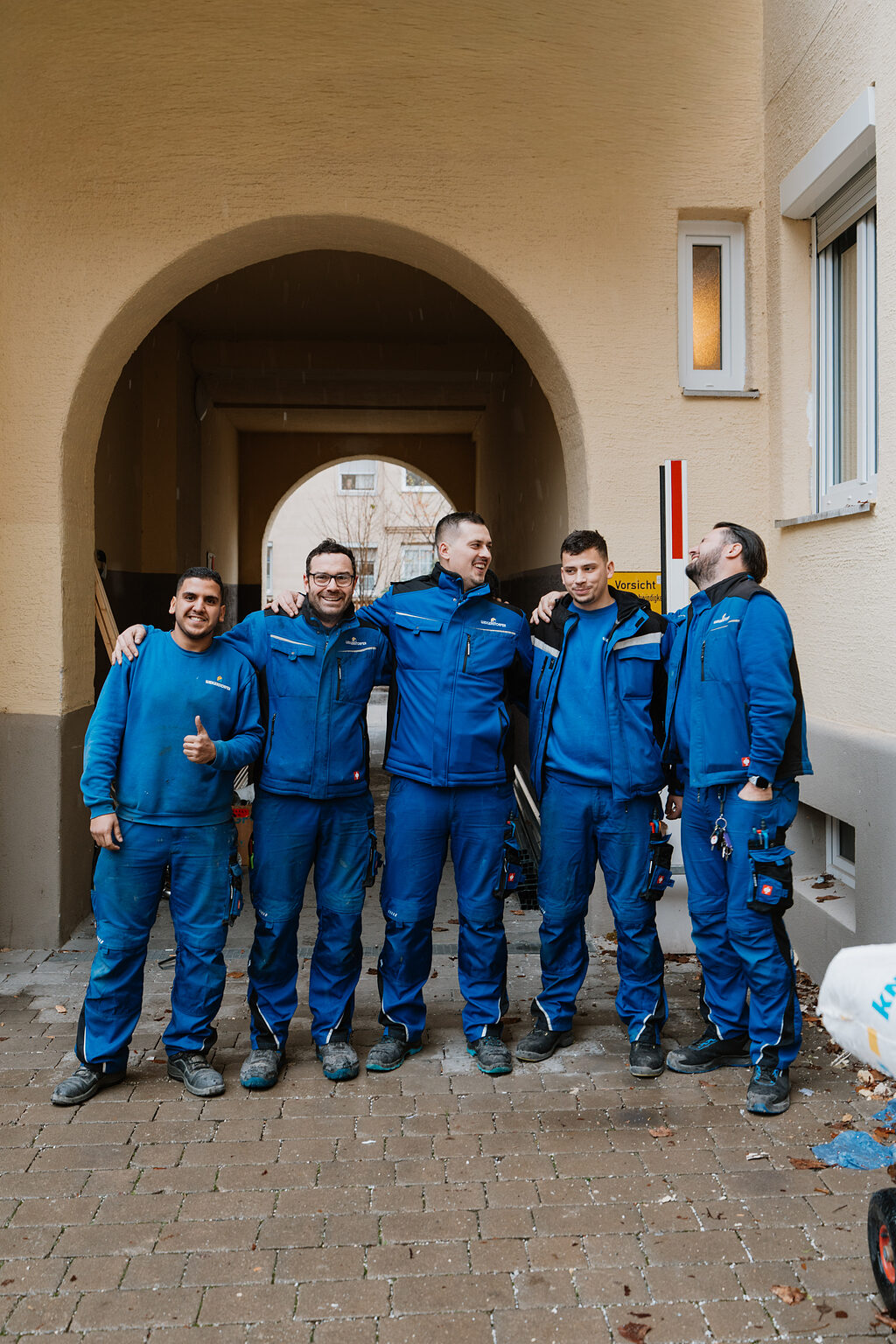 Fünf Handwerker der Weigerstorfer GmbH in blauer Arbeitskleidung stehen nebeneinander vor einer Baustelle